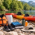 Chaleco, calzado cerrado, ropa, agua y bidón estanco junto a un kayak doble en un río